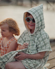 Child wearing a green checkered poncho sitting by a body of water with another child in the background.