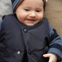 Baby wearing a navy coat and striped hat with a brown interior, smiling.