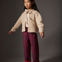 Young girl wearing a cream sweater with a bow and patterned burgundy pants on a gray background