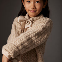 Young girl wearing a beige cable knit sweater with a bow tie against a gray background