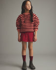 A young girl stands against a neutral gray studio background wearing a red and pink striped knit sweater, a red corduroy mini skirt, dark red socks, and dark gray boots.