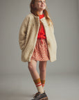 A young girl with red hair stands against a neutral gray studio background wearing a cream sherpa fleece jacket over a bright red top, a red and white patterned skirt, striped socks, and dark brown shoes.