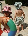 Two children wearing colorful hats and swimwear, one holding a life buoy, on a sunlit patio.