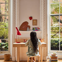 Child sitting at a desk in a bright room with large windows, surrounded by books and a red lamp.