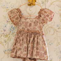 Floral dress with ruffled sleeves and bottom hanging on a decorative gold hanger against a floral wallpaper background.