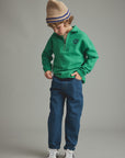 A child wearing a bright green half-zip pullover, blue jeans, white sneakers, and a beige knit beanie with blue stripes, standing against a plain gray studio background.