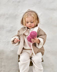 A young child wearing a beige hooded jacket with cream sherpa lining and cream pants, sitting on a wooden stool against a textured light gray wall and holding a ball of pink yarn.