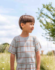 Child wearing a checkered outfit standing in a grassy field with a blue sky.