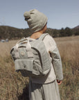 A child wearing a beige knit beanie and matching outfit stands in a dry grassy field, viewed from behind while wearing a light grayish-beige backpack.