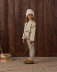 A young girl stands in front of a dark wood plank wall wearing a cream knit beanie, a cream patterned jacket with dark trim, light beige leggings, and tan slippers. A stack of books and part of a wooden chair are visible on the left.