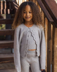 A young girl with curly hair stands on wooden stairs wearing a light gray knit cardigan and matching ribbed pants.