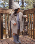 A young girl stands on a wooden deck wearing a long beige coat, a cream knit beanie, a blue button-up shirt, a gray skirt, and gray boots, while holding a vintage-style lantern. Trees and a cabin are visible in the background.