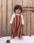 A toddler stands in snow in front of a wooden fence, wearing rust and tan vertically striped overalls over a cream long-sleeve shirt and tan boots.