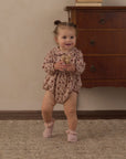A toddler girl wearing a long-sleeve floral romper and pink booties is standing and smiling in a room with a wooden dresser and beige wall.