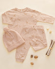 A baby clothing set laid flat on a warm cream background, including a long-sleeve top, pants, and a knit beanie with a pom-pom. The set is light blush pink with a subtle deer print and small polka dots, styled with natural props like pinecones, acorns, and twigs.