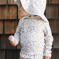 Child wearing a floral sun hat and swimsuit against a wooden wall.