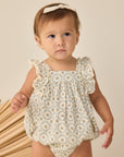 Baby wearing a floral romper standing next to wooden sticks on a beige background