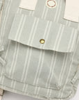 Close-up of a canvas backpack in a pale sage green and white stripe pattern, featuring a front pocket with a brass snap button, off-white webbing straps, and a circular brand patch reading 'MILKY CAKE TOYS'.