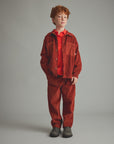 A young boy with red hair stands against a neutral gray studio background wearing a rust-orange corduroy jacket and matching corduroy pants over a bright red collared shirt, with dark gray boots.