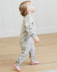Child wearing a tie-dye onesie standing on a wooden floor.