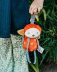 Child holding a small plush toy with an orange outfit and red hat against a natural background.