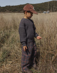 A child standing in a dry grassy field wearing a dark gray cable-knit sweater, plaid pants, and a brown corduroy cap.