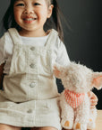 Child in a white dress with a beige checkered apron holding a plush toy on a wooden surface.