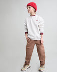 A child model wearing a white crewneck sweatshirt with burgundy trim, brown pants, a red knit beanie, and patterned sneakers, standing against a solid light gray background.