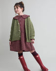 Child wearing a green cardigan, patterned dress, and red boots on a plain background