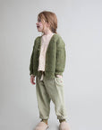 A young girl wearing a green chunky knit cardigan over a cream polka dot top, sage green pants, and fuzzy beige slippers, standing against a light gray studio background.