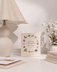 Book titled 'Our Little Library' on a table with a lamp and flowers in the background
