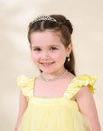 A young girl with brown hair in a half-up style wearing a silver tiara, matching silver necklace, and a yellow dress with ruffled shoulders, photographed against a soft beige studio background.