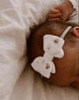 Baby wearing a white headband with a bow, lying on a soft beige blanket.