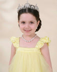 A young girl with dark hair styled in a braid, wearing a silver tiara, a pearl necklace, and a pale yellow dress with ruffled shoulders, set against a soft beige background.