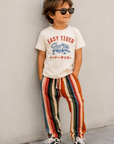 Child wearing a 'Easy Tiger' t-shirt and colorful striped pants against a plain wall.