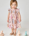 Young girl wearing a pink dress with rabbit pattern, standing against a plain background with toy rabbits around.