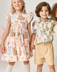 Three children wearing floral and patterned outfits with decorative flowers in the background.
