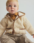 A baby wearing a cream sherpa fleece hooded jacket with brown trim, a mustard yellow shirt, and brown and beige striped pants, set against a plain light gray background.