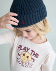 A child wearing a dark navy blue ribbed knit beanie with a small embroidered red logo and a cream graphic t-shirt, photographed against a light gray background.