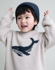 Child wearing a sweater with a whale design and a knit hat on a plain background