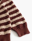 A close-up of a striped knit sweater sleeve with alternating horizontal bands of deep burgundy and warm beige, featuring textured cable-knit and diamond-patterned sections with ribbed cuffs.