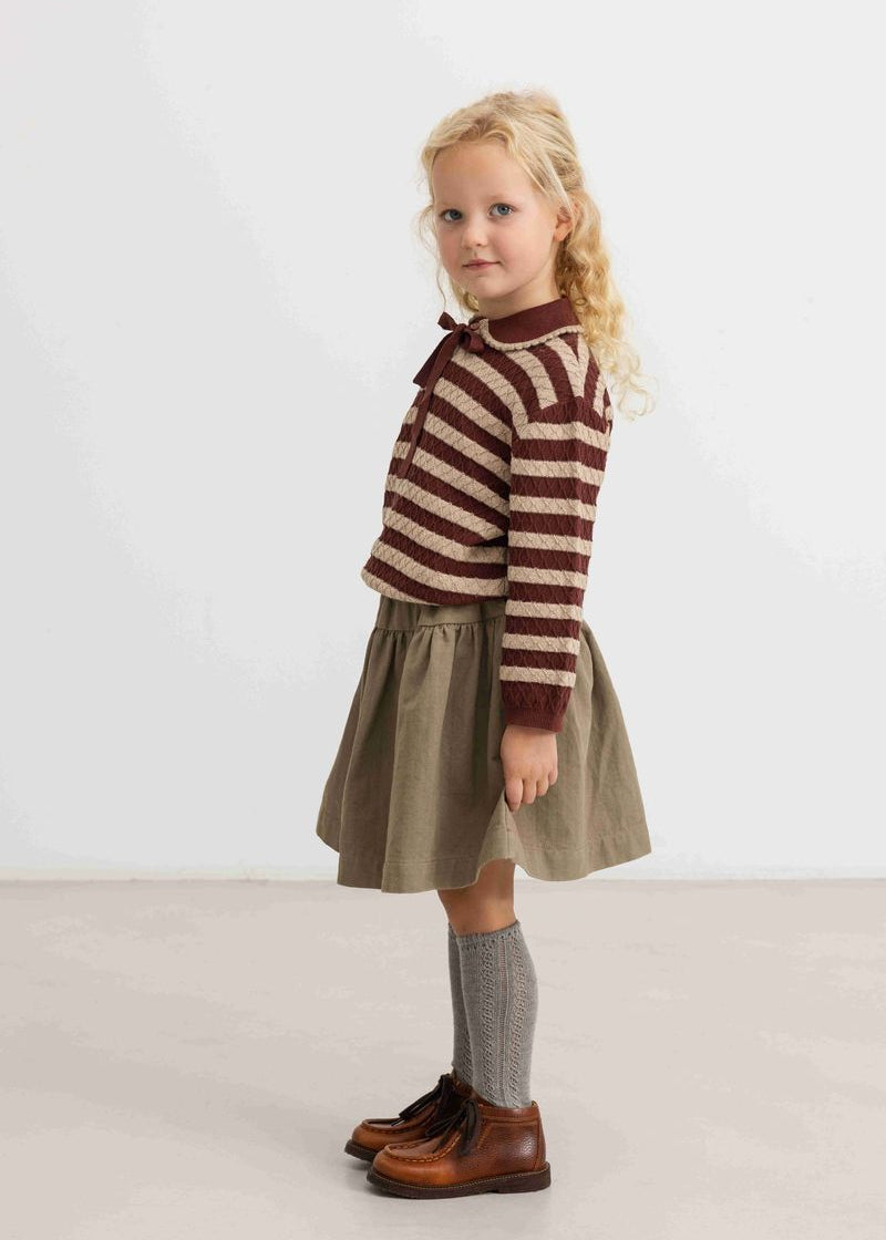A young girl with blonde curly hair stands in profile wearing a brown and beige striped knit sweater, an olive green skirt, gray knee-high socks, and brown leather shoes against a plain light gray studio background.