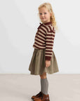 A young girl with blonde curly hair stands in profile wearing a brown and beige striped knit sweater, an olive green skirt, gray knee-high socks, and brown leather shoes against a plain light gray studio background.