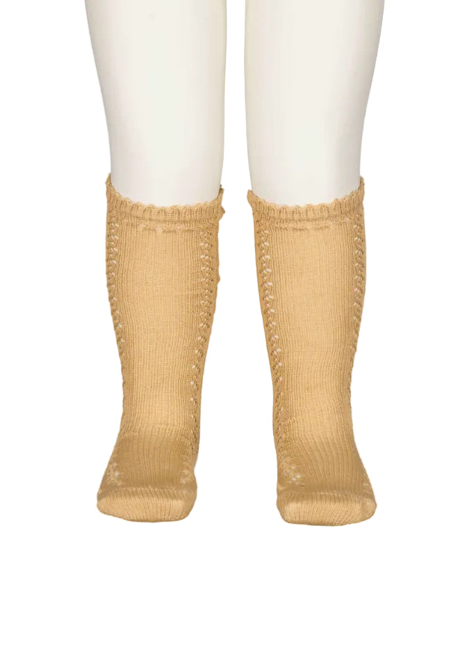 A pair of beige ribbed knit socks with decorative openwork lace-like patterns along the sides and scalloped ruffle cuffs, shown on a white mannequin lower leg against a white background.