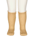 A pair of beige ribbed knit socks with decorative openwork lace-like patterns along the sides and scalloped ruffle cuffs, shown on a white mannequin lower leg against a white background.
