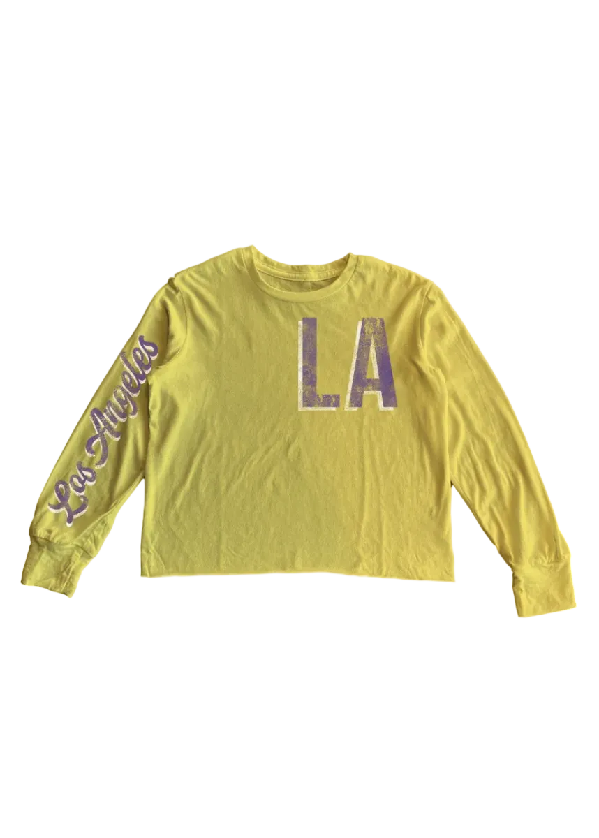 A yellow long-sleeve crop top featuring 'Los Angeles' in purple script on the right sleeve and distressed purple 'LA' block letters on the center chest, displayed flat against a white background.