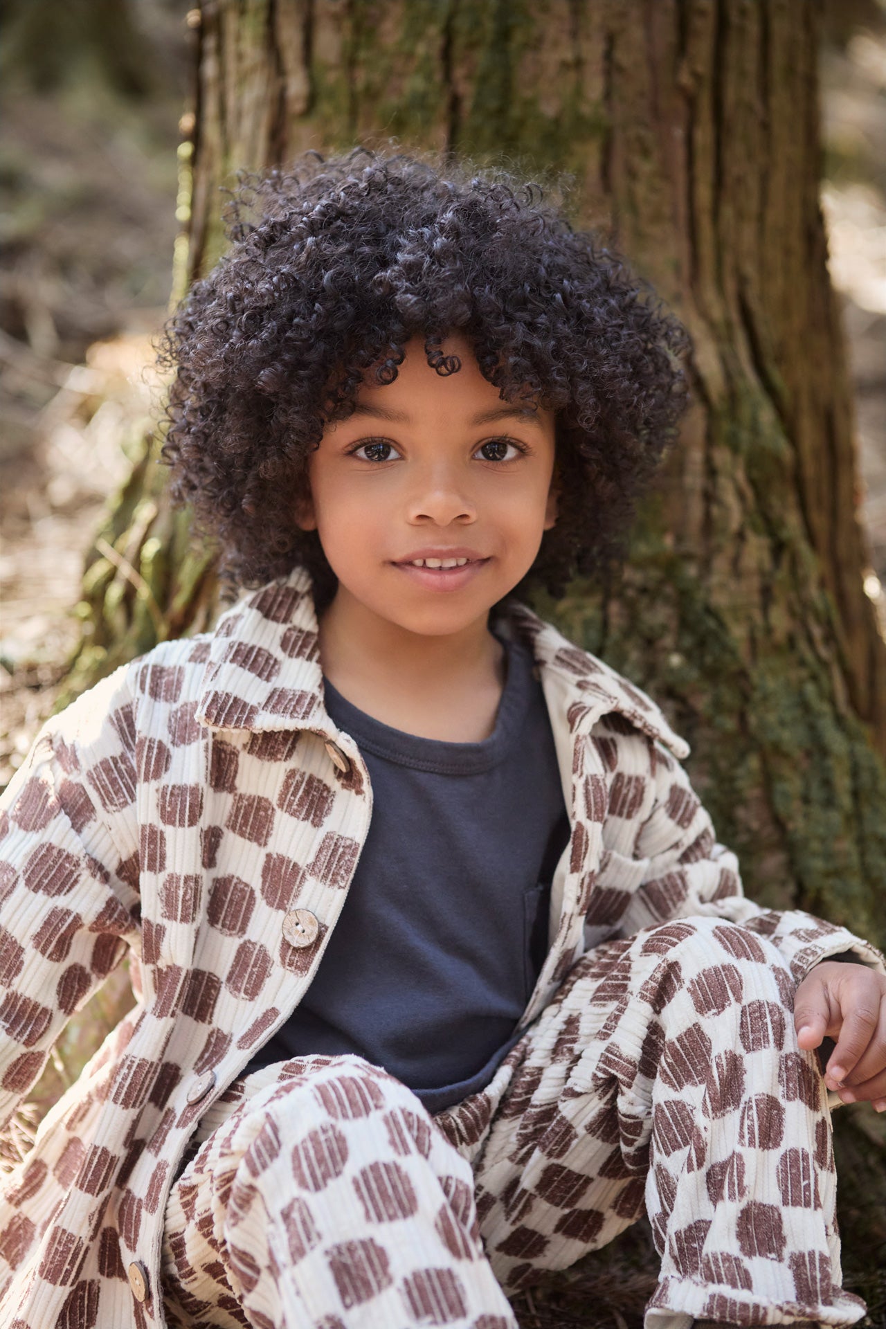 Child sitting outdoors in front of a tree wearing a patterned coat.