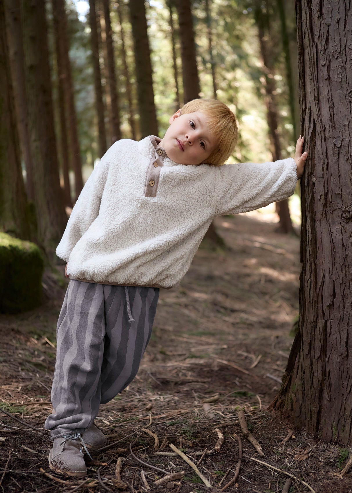 Child in a white sweater and gray pants standing next to a tree in a forest