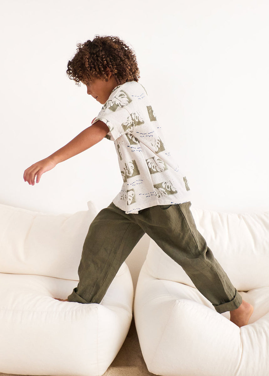 Child in a white shirt with a map design and green pants standing on a white couch.