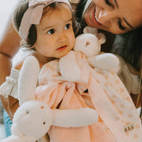 Woman holding a baby dressed in a pink outfit with a bow, surrounded by soft toys.